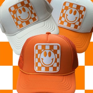 May include: Three trucker hats with orange and white checkered smiley face patches. The hats are white, orange, and orange.