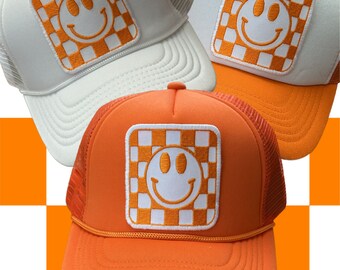 May include: Three trucker hats with orange and white checkered smiley face patches. The hats are white, orange, and orange.