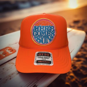 May include: Orange trucker hat with a circular patch that says "Here Comes The Sun" in a retro font. The patch has a pink, yellow, and blue color scheme. The hat has a black label that says "OTTO World Famous Foam Front OSFM Trucker Durable Snap Closure".