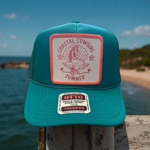 May include: A turquoise trucker hat with a white patch featuring a mermaid sitting on a seashell with the text "Coastal Cowgirl Summer". The hat has a black mesh back and a black adjustable snap closure.