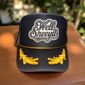 May include: Black trucker hat with the text "Well Sheeyit" in white script. The hat features gold embroidered accents on the front and a gold rope detail. The hat has a mesh back.