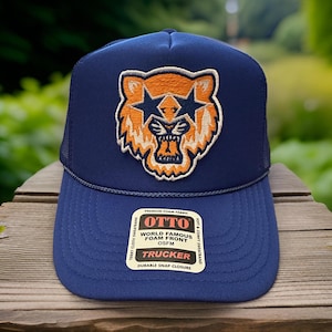 May include: A blue trucker hat with a foam front and a large orange tiger with star-shaped eyes embroidered on the front. The hat has a black mesh back and a white label with the text "OTTO WORLD FAMOUS FOAM FRONT OSFM TRUCKER DURABLE SNAP CLOSURE".