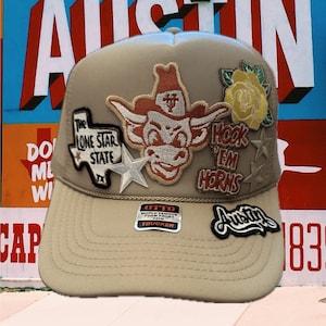 May include: A beige trucker hat with a variety of patches, including a Longhorn steer, a star, a rose, and a "Hook 'Em Horns" patch. The hat also has a black and white "Otto" label and a black and white "Austin" patch.