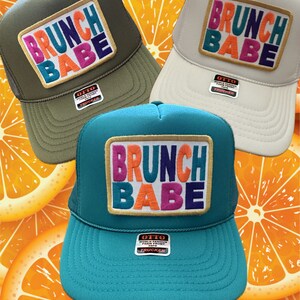 May include: Three trucker hats with the text "BRUNCH BABE" in colorful embroidered letters. The hats are in olive green, cream, and teal. Each hat has a foam front and a mesh back. The background features orange slices.