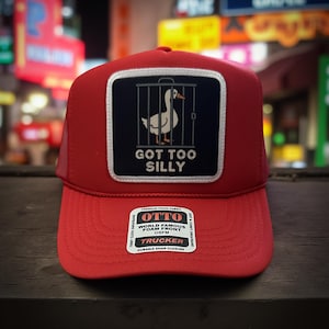 May include: Red trucker hat with a white and black patch featuring a goose in a cage and the text "GOT TOO SILLY." The hat has a mesh back and a curved brim. The hat also has a white sticker with the text "OTTO TRUCKER".