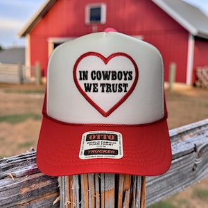 May include: A red and white trucker hat with the text "IN COWBOYS WE TRUST" inside a heart. The hat has a red brim and a white front panel. The hat is made by Otto and has a durable snap closure.