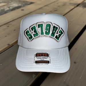 May include: White trucker hat with the word "EAGLES" in green, arched lettering. The hat has a white foam front and a mesh back. The hat includes a tag that says "OTTO WORLD FAMOUS FOAM FRONT OSFM TRUCKER DURABLE SNAP CLOSURE".