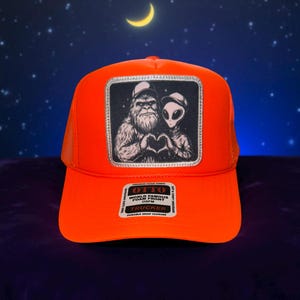 May include: Bright orange trucker hat with a mesh back and a patch featuring a Bigfoot and an alien forming a heart shape. The patch is bordered in silver. The hat has the text "OTTO WORLD FAMOUS FOAM FRONT TRUCKER" on a tag.