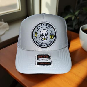 May include: A white trucker hat with a mesh back and a curved brim. The front features a circular patch with a skull, lightning bolt, and coffee cup, and the text "NO TALKING BEFORE COFFEE".