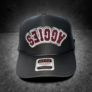 May include: Black trucker hat with the word "AGGIES" in maroon and white lettering. The hat has a mesh back and a curved brim. The hat has a tag that says "OTTO WORLD FAMOUS FOAM FRONT OSFM TRUCKER DURABLE SNAP CLOSURE".