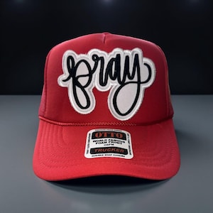 May include: Red trucker hat with the word "pray" in white script outlined in black. The hat has a mesh back and a curved brim. The front of the hat features a foam front and a durable snap closure.