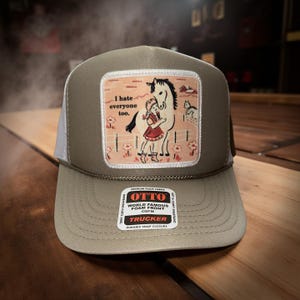 May include: A trucker hat with a tan front panel and white mesh back. The hat features a patch with a girl hugging a horse, with the text "I hate everyone too." The hat has a curved brim and a snap closure.