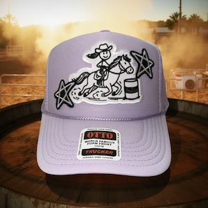 May include: A lavender trucker hat with a white mesh back and a curved brim. The front features a patch of a stick figure riding a horse around a barrel, with star accents. The hat has the text "OTTO WORLD FAMOUS FOAM FRONT TRUCKER".
