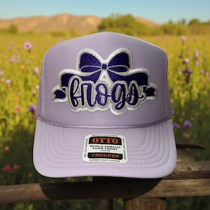 May include: A lavender trucker hat with a white brim and mesh back. The hat features the word "frogs" in a cursive font with a bow design above it. The hat has a snap closure and a "OTTO" label.