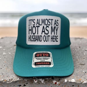 May include: A teal trucker hat featuring a white rectangular patch with black embroidered text that reads, IT'S ALMOST AS HOT AS MY HUSBAND OUT HERE. The hat includes a sticker on the brim with the brand name OTTO and product details.