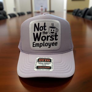 May include: A lavender trucker hat with a white patch that reads "Not the Worst Employee" with a coffee cup graphic. The hat has a curved brim and a mesh back. The brand name "OTTO" is visible on a tag.
