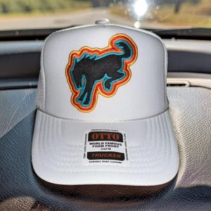 May include: A white trucker hat featuring a black bucking horse patch outlined in blue, orange, and yellow. The front panel has a sticker that reads OTTO WORLD FAMOUS FOAM FRONT OSFM TRUCKER, TERRY CLOTH SWEATBAND, SOFT & COMFY SWEATBAND, DURABLE SNAP CLOSURE.