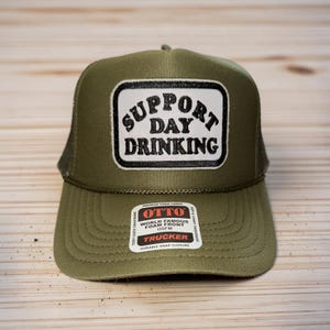May include: Olive green trucker hat with a white and black patch that reads "SUPPORT DAY DRINKING." The hat has a mesh back and a curved brim. The hat is made of foam front fabric and has a durable snap closure.