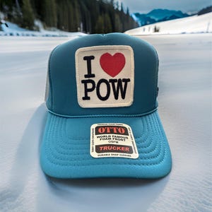 May include: A blue trucker hat with a white patch that says "I love POW" with a red heart. The hat has a mesh back and a snap closure.