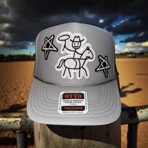 May include: A gray trucker hat with a mesh back and a foam front. The front features a white embroidered design of a cowboy riding a horse, with two black star accents. The hat has a snap closure and a tag that reads "OTTO".