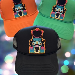 May include: Three trucker hats with a colorful embroidered nutcracker design. The hats are orange, green, and black.