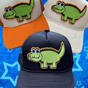 May include: Three trucker hats with a green alligator wearing sunglasses embroidered on the front. The hats are white, orange, and navy blue.