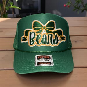 May include: Green trucker hat with the word "Bears" in white and gold script, accented by a green bow. The hat features a mesh back and a curved brim. The hat has a "World Famous Foam Front" tag.