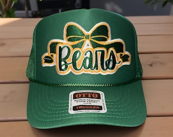 May include: Green trucker hat with the word "Bears" in white and gold script, accented by a green bow. The hat features a mesh back and a curved brim. The hat has a "World Famous Foam Front" tag.