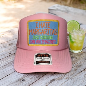 May include: Pink trucker hat with a rectangular patch that reads "I HATE MARGARITAS JUST KIDDING CAN YOU IMAGINE?" in embroidered text. A margarita cocktail with a lime wedge sits next to the hat. The hat has a snap closure.