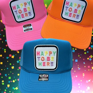 May include: Three trucker hats with a square patch that says "Happy to be here" in rainbow colors. The hats are pink, orange, and blue.