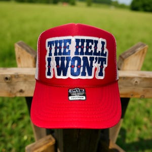 May include: A red trucker hat with a white mesh back. The front of the hat features the text "THE HELL I WON'T" in white and blue lettering. The hat has a curved brim and a small tag that says "OTTO".