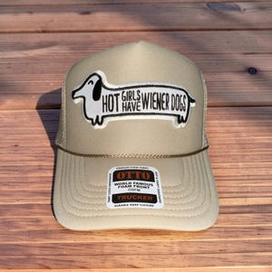 May include: Tan trucker hat with a white and black wiener dog graphic and the text "HOT GIRLS HAVE WIENER DOGS." The hat has a foam front and a mesh back. The hat also has a snap closure.