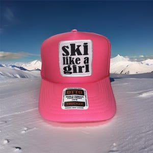 May include: Pink trucker hat with a black and white patch that says "ski like a girl". The hat has a black mesh back and a black label that says "OTTO World Famous Foam Front OSFM Trucker Durable Snap Closure".