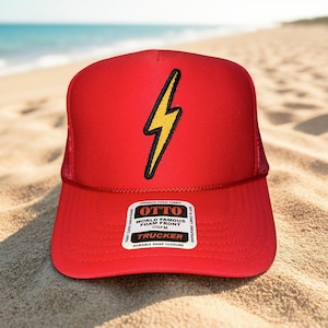 May include: A red trucker hat with a yellow and black lightning bolt graphic. The hat has a foam front and a mesh back. The hat has the text "OTTO WORLD FAMOUS FOAM FRONT TRUCKER" on a white label.