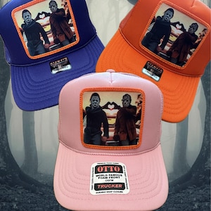 May include: Three trucker hats with a square patch featuring a horror movie character. The hats are blue, orange, and pink. The patch shows two characters making a heart shape with their hands. The hats have the text "OTTO WORLD FAMOUS FOAM FRONT TRUCKER" on the front.