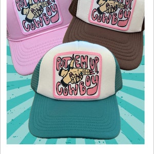 May include: Three trucker hats with a pink and white patch that says "Put 'em Up Cowboy" in a pink and white font. The hats are pink, brown, and teal.