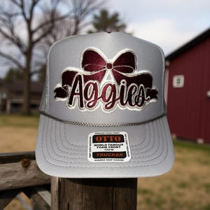 May include: Gray trucker hat with a maroon bow and the word "Aggies" embroidered on the front. The hat features a mesh back and a snap closure. The hat is made of premium foam fabric and has a terry cloth sweatband.