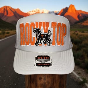 May include: White trucker hat with the text "ROCKY TOP" in orange and a black dog graphic. The hat has a white front panel, a white brim, and a white mesh back. The hat has a tag that says "OTTO WORLD FAMOUS FOAM FRONT TRUCKER".