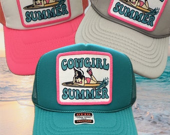 May include: Three trucker hats with a pink, beige, and teal colored mesh back. Each hat has a square patch with a cowgirl on a surfboard and the text "Cowgirl Summer". The teal hat has a small black and white label that says "Otto World Famous Foam Front Trucker".