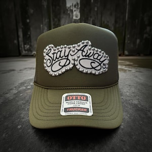 May include: Olive green trucker hat with the text "Stay Away" in a stylized font, embellished with silver studs. The hat features a mesh back and a curved brim. The label reads "OTTO World Famous Foam Front OSFM Trucker".