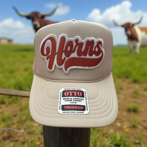 May include: A beige trucker hat with a red and white "Horns" logo. The hat has a mesh back and a foam front. The hat is sitting on a wooden fence post.