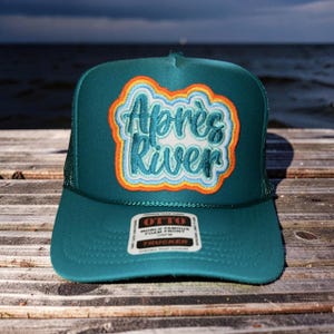 May include: Teal trucker hat with the text "Après River" in a colorful, embroidered patch. The hat has a mesh back and a curved brim. A white tag with black text is attached to the front.