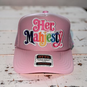 May include: A pink trucker hat with the text "Her Majesty" in colorful, embroidered lettering. The hat features a mesh back and a curved brim. The front of the hat has a patch that reads "OTTO World Famous Foam Front".