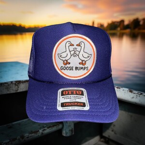 May include: A navy blue trucker hat with a white and orange circular patch featuring two cartoon geese and the words "GOOSE BUMPS". The hat has a mesh back and a snap closure. The brand name "OTTO" is visible.