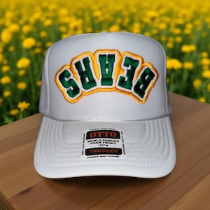 May include: White trucker hat with the word "BEARS" in green and gold lettering across the front. The hat has a foam front and a durable snap closure. The hat is made of a soft and comfortable material.