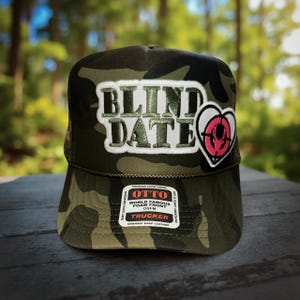 May include: A camouflage trucker hat with the text "BLIND DATE" in green letters on a white patch. A pink and white heart-shaped patch with a target design is on the right side. The hat has a mesh back and a snap closure.