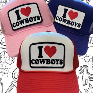 May include: Three trucker hats with a red heart and the text "I Love Cowboys" on a white patch. The hats are pink, blue, and red.