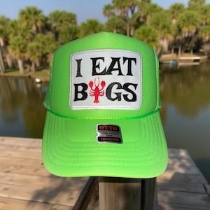 May include: A bright green trucker hat with a white patch that says "I EAT BUGS" in black letters with a red lobster graphic. The hat has a black mesh back and a black "OTTO WORLD FAMOUS TRUCKER CAP" label.