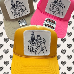 May include: Three trucker hats with a black and white graphic of two men with beards. The hats are in beige, pink, and yellow.