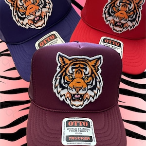 May include: Three trucker hats with a tiger face design. The hats are navy blue, burgundy, and red. The hats have a mesh back and a foam front. The hats have a label that says "OTTO World Famous Foam Front OSFM Trucker Durable Snap Closure".